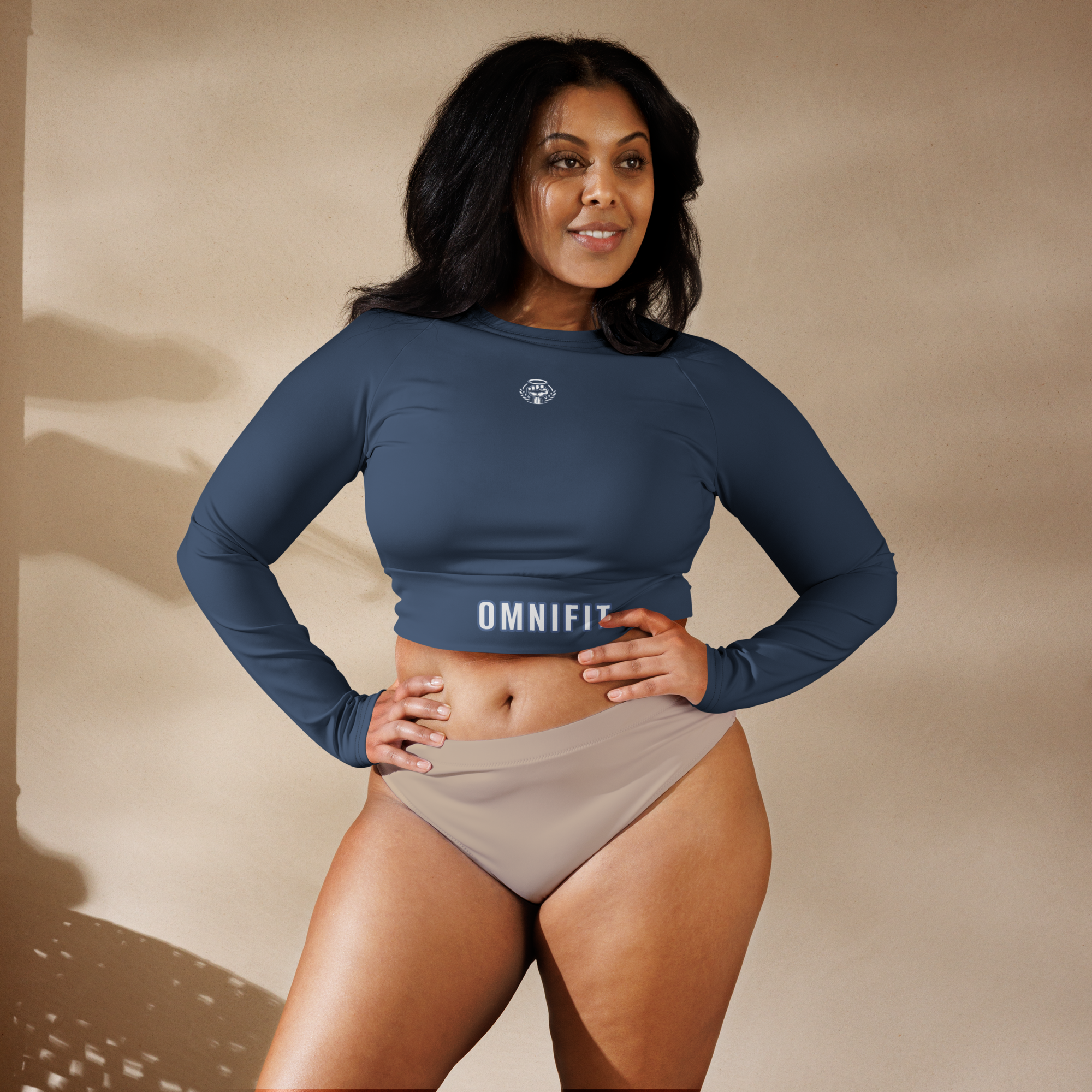 Woman Posing in OmniFit Activewear A confident woman with dark hair poses in a studio setting, wearing a long-sleeved, dark blue cropped top from OmniFit and beige bottoms.