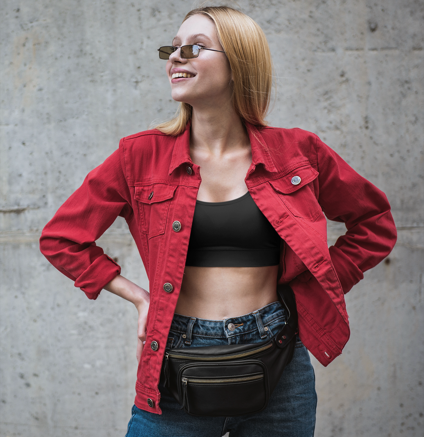 Stylish Woman in Red Jacket and Sports Bra A fashionable woman with blonde hair is smiling confidently, wearing a black sports bra, a red denim jacket, and blue jeans with a black fanny pack.