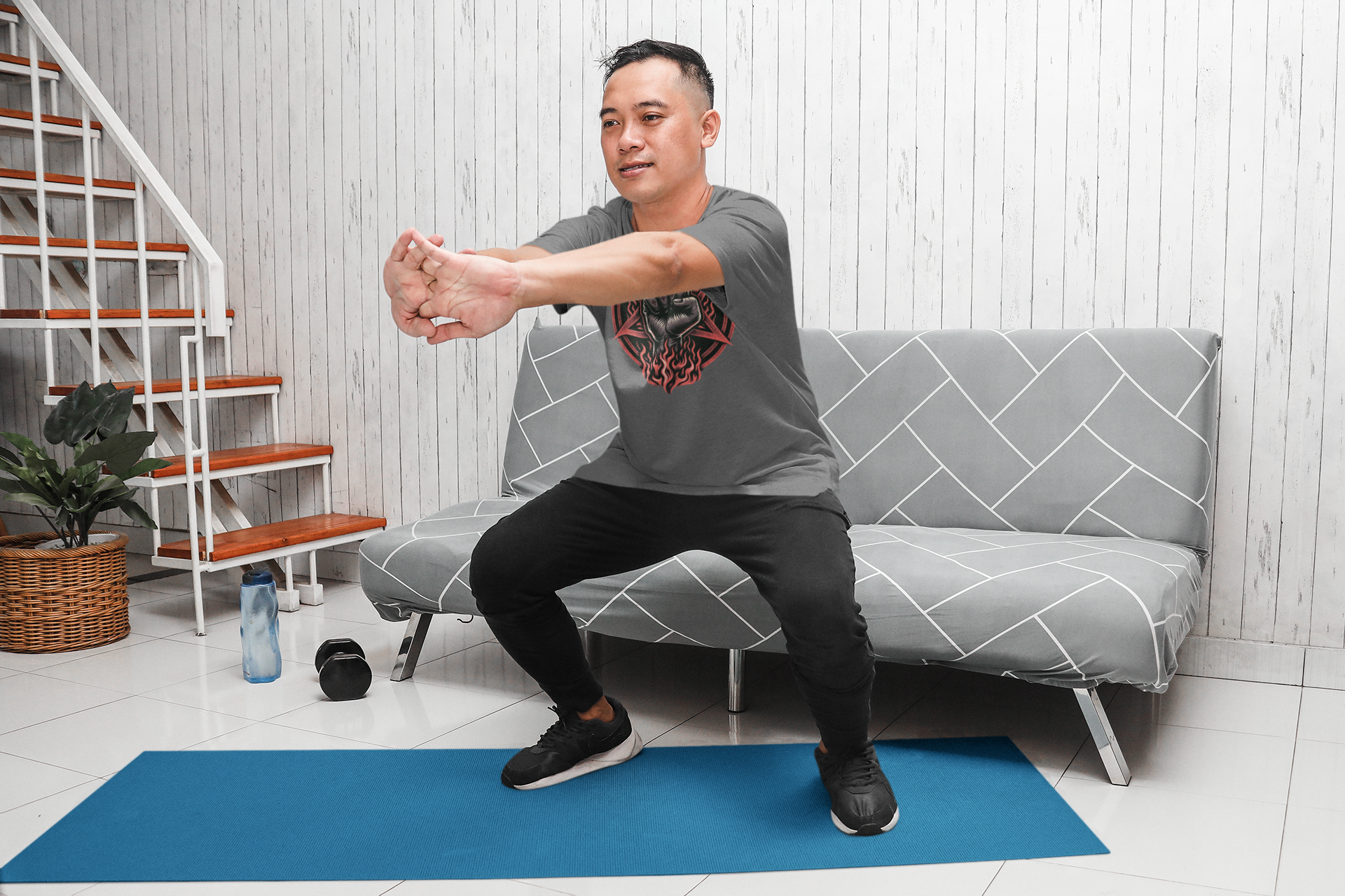Man Exercising at Home on a Yoga Mat A man is doing a squat exercise on a blue yoga mat in his living room. He is wearing a gray graphic t-shirt and black pants, with a water bottle and dumbbell placed nearby.