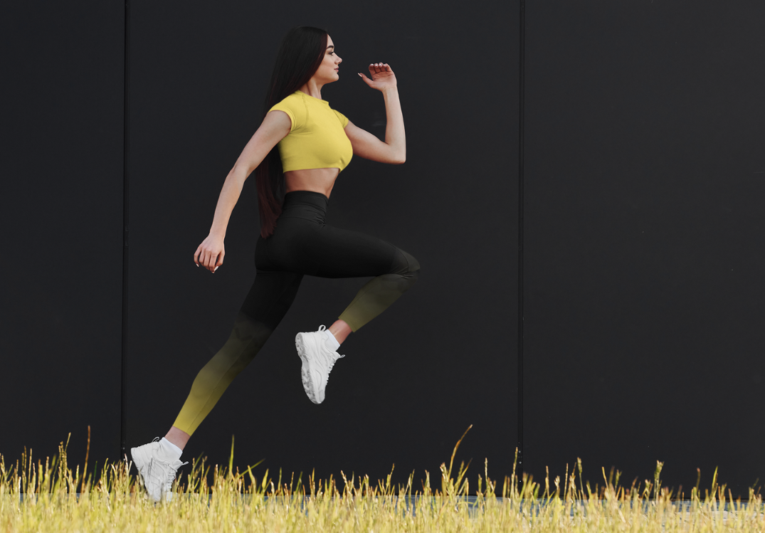 Woman Running in OmniFit Activewear A brunette woman with long hair is captured mid-stride as she runs against a dark background. She is wearing a yellow cropped top and black leggings from OmniFit, along with white athletic shoes.