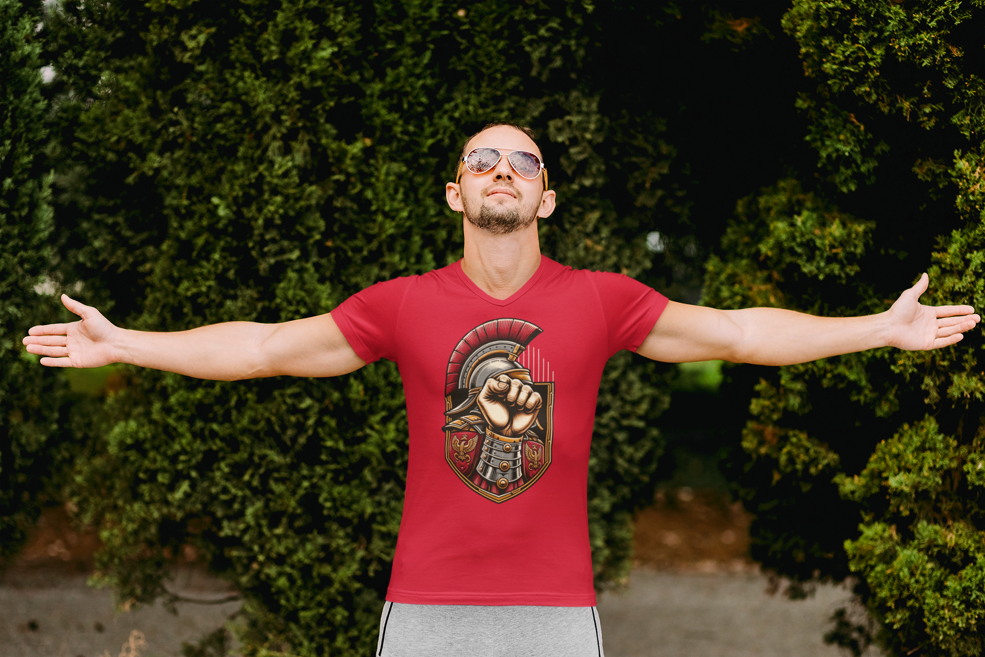 Man with Open Arms in Red Graphic Tee A man wearing sunglasses and a red V-neck t-shirt with a graphic of a fist inside a Roman helmet is standing with his arms outstretched.